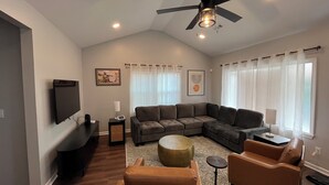 A living room with a sectional sofa, a flat-screen TV, a ceiling fan, and a large window with sheer curtains.