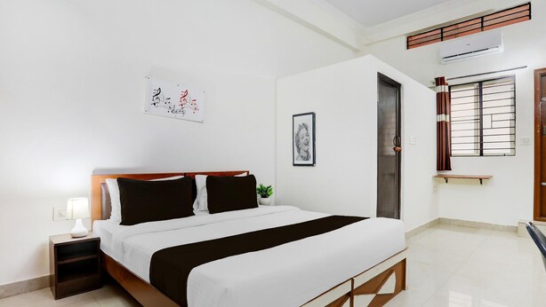A bedroom with a bed, bedside table, lamp, wall art, and a window with a view. - Deluxe Sri Krishna Nilaya (Bengaluru)