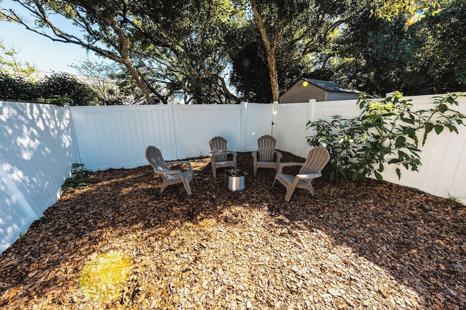 Ein Garten mit weißem Zaun, vier Adirondack-Stühlen, einer Feuerstelle aus Metall und einem Baum.