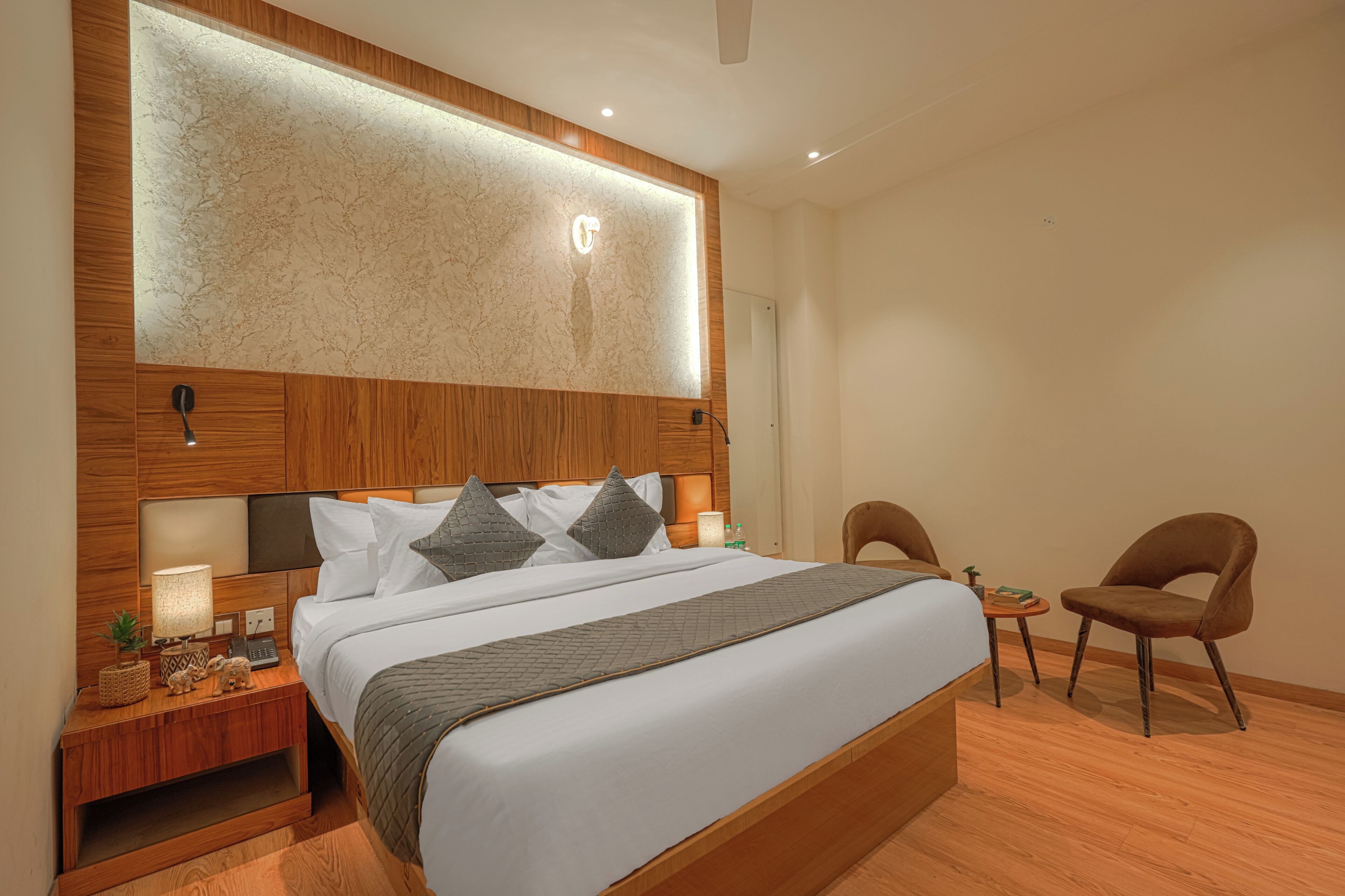 A modern hotel room with a large bed, wooden headboard, bedside tables, a small desk, and a chair.
