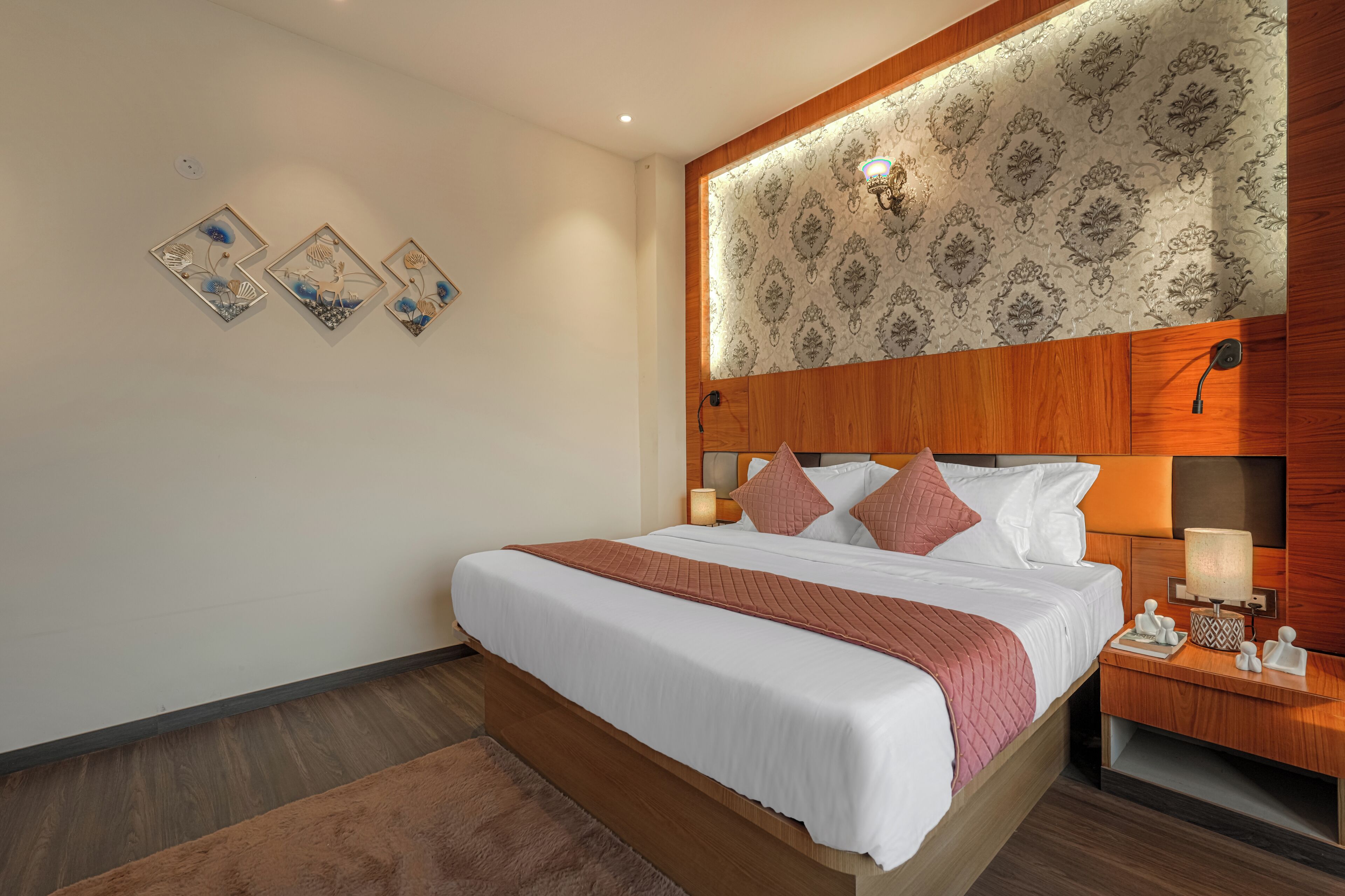 Une chambre d’hôtel avec une tête de lit en bois, un lit recouvert de draps blancs, une table de chevet avec une lampe et trois œuvres d’art accrochées au mur.