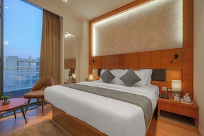 A modern hotel room with a large bed, a wooden headboard, bedside lamps, a nightstand, and a view of the city. - Hotel Aurelia Inn Near Airport Road Sector 82 Mohali Chandigarh (Mohali)