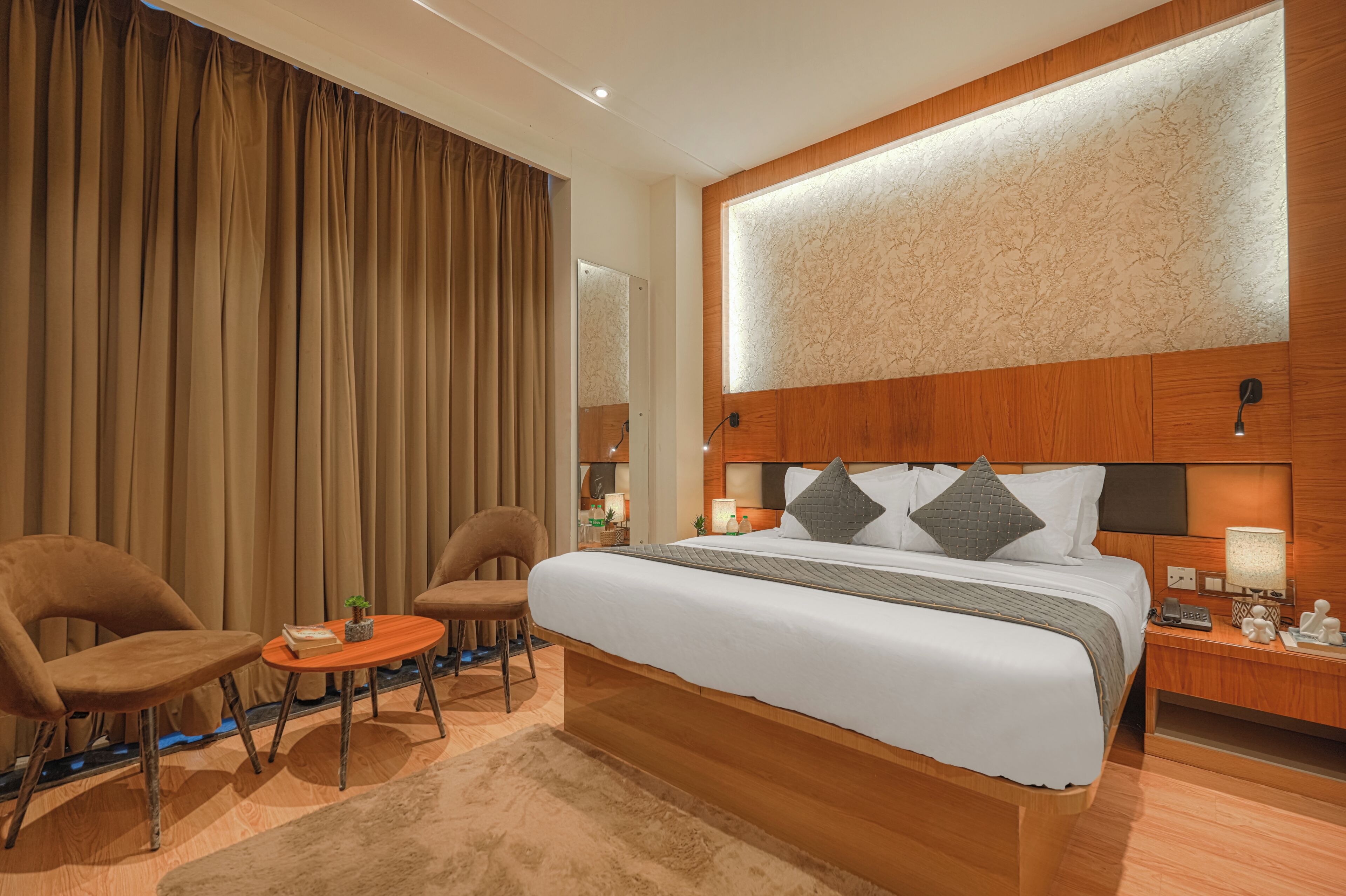 A hotel room with a large bed, two armchairs, a small table, and a nightstand with a lamp.