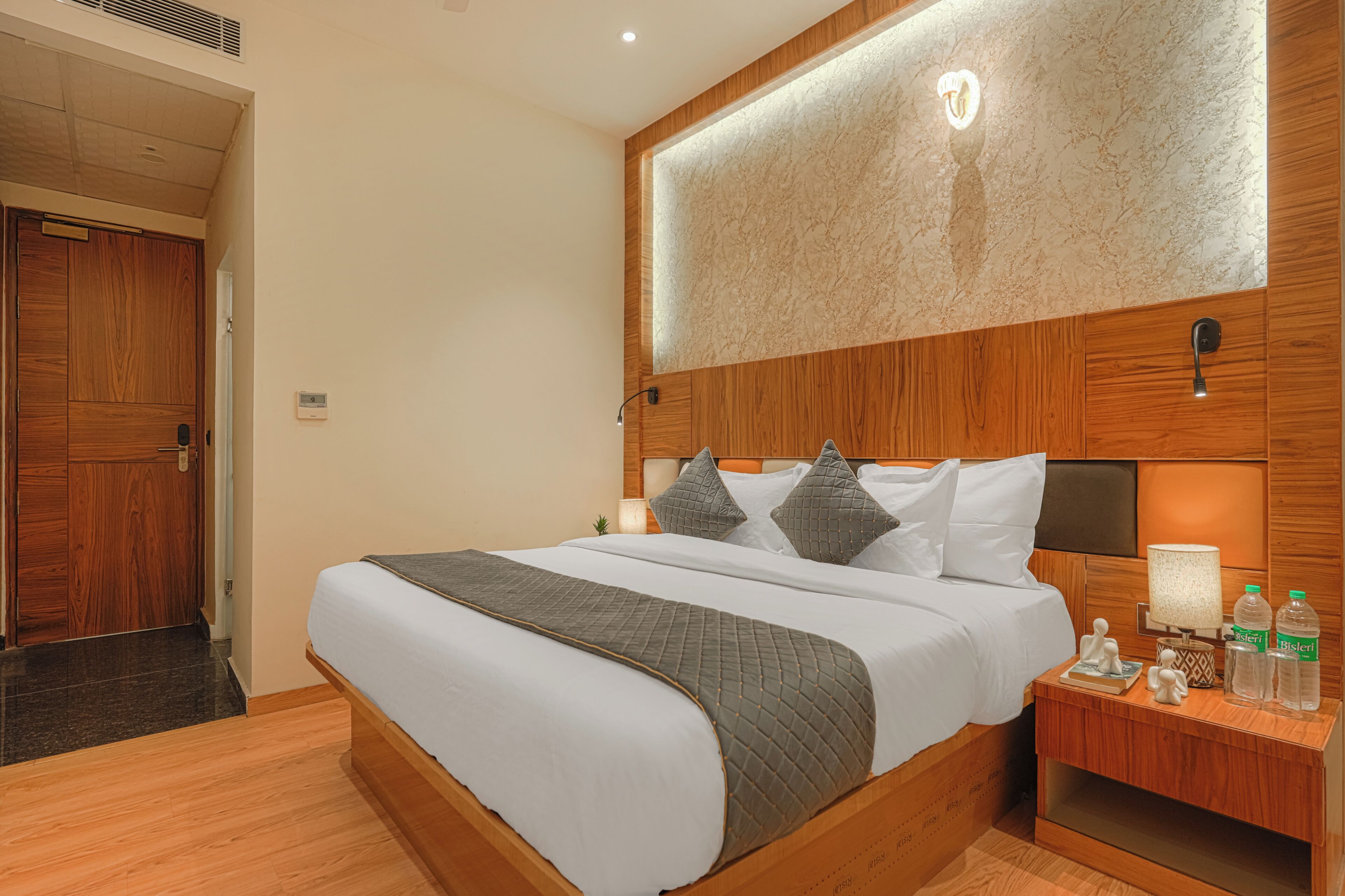 A hotel room with a wooden bed, a nightstand with a lamp, and a wall-mounted light fixture.