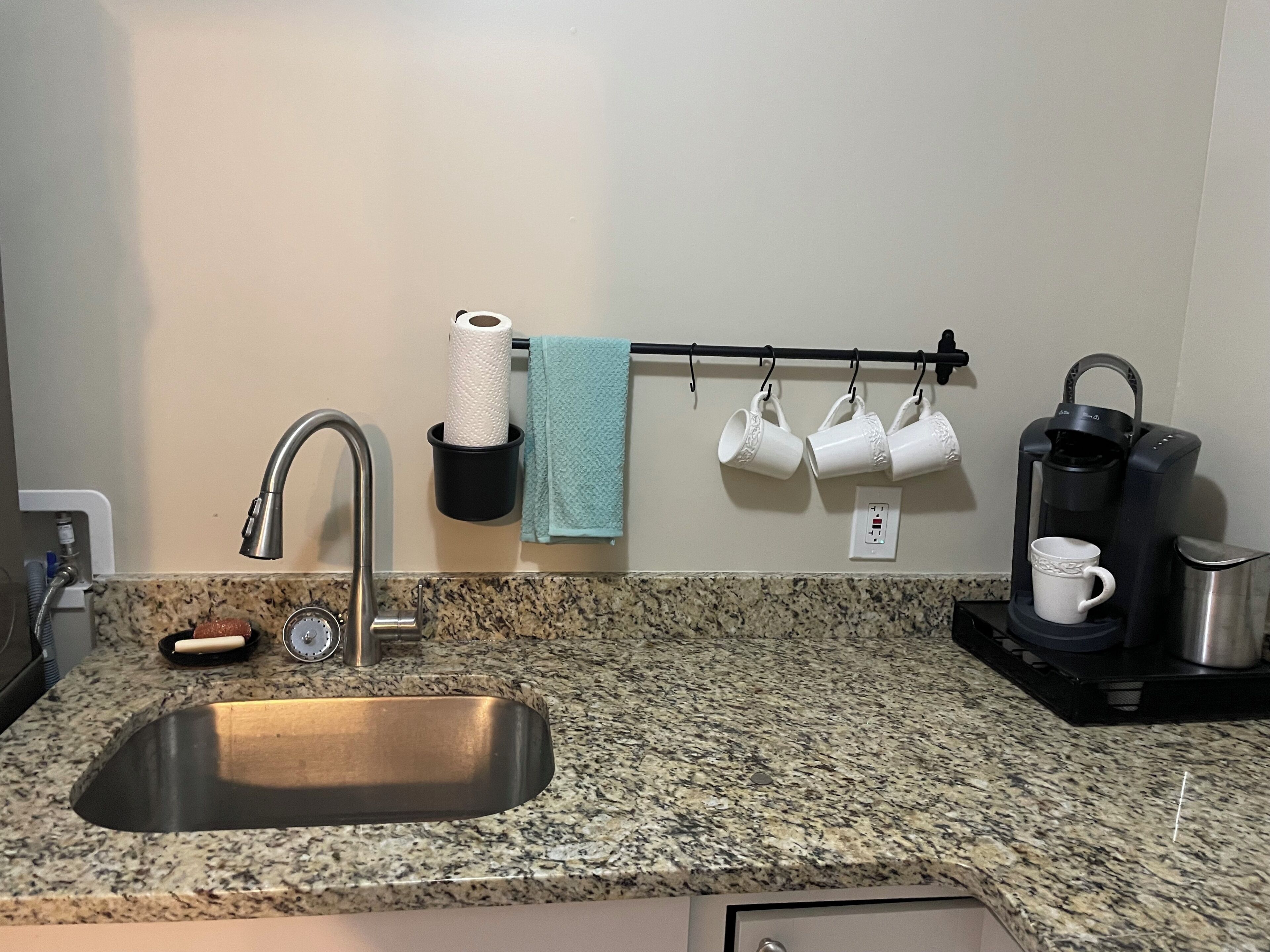Eine Küche mit Granitarbeitsplatte, einer Edelstahlspüle, einer Kaffeemaschine und einem Handtuchhalter mit Tassen.