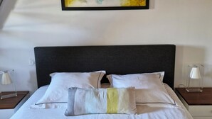 Un lit recouvert d’une courtepointe matelassée, avec une tête de lit noire et des oreillers blancs. Au-dessus du lit, on aperçoit un tableau abstrait encadré, et une table de chevet avec une lampe se trouve sur le côté droit.