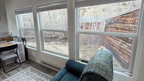 A room with a blue sofa, a desk with a chair, and large windows with a view of a wooden fence and trees.