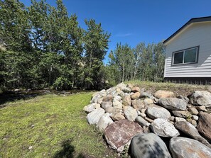 A house with a stone retaining wall, a grassy yard, and dense trees in the background. - Unique creek side, pet friendly tiny home on the outskirts of Sheridan MT. (Sheridan)