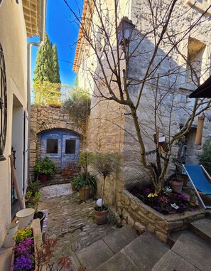 Un patio estrecho con muros de piedra, un pequeño jardín con plantas en macetas, una puerta azul y una escalera de piedra.