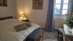 Ein Schlafzimmer mit Bett, Nachttischlampe, Kunstwerk, Fenster mit Vorhängen und einem kleinen Tisch mit Blumen.