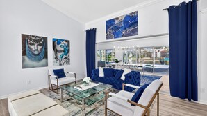 A spacious living room with a glass coffee table, a blue sofa, and a white armchair. There are two large paintings on the wall, a chandelier, and a view of the outside. - Blu Breeze, Social Media moments, Volleyball court, 4 BD, Largest Canal front. (Cape Coral)