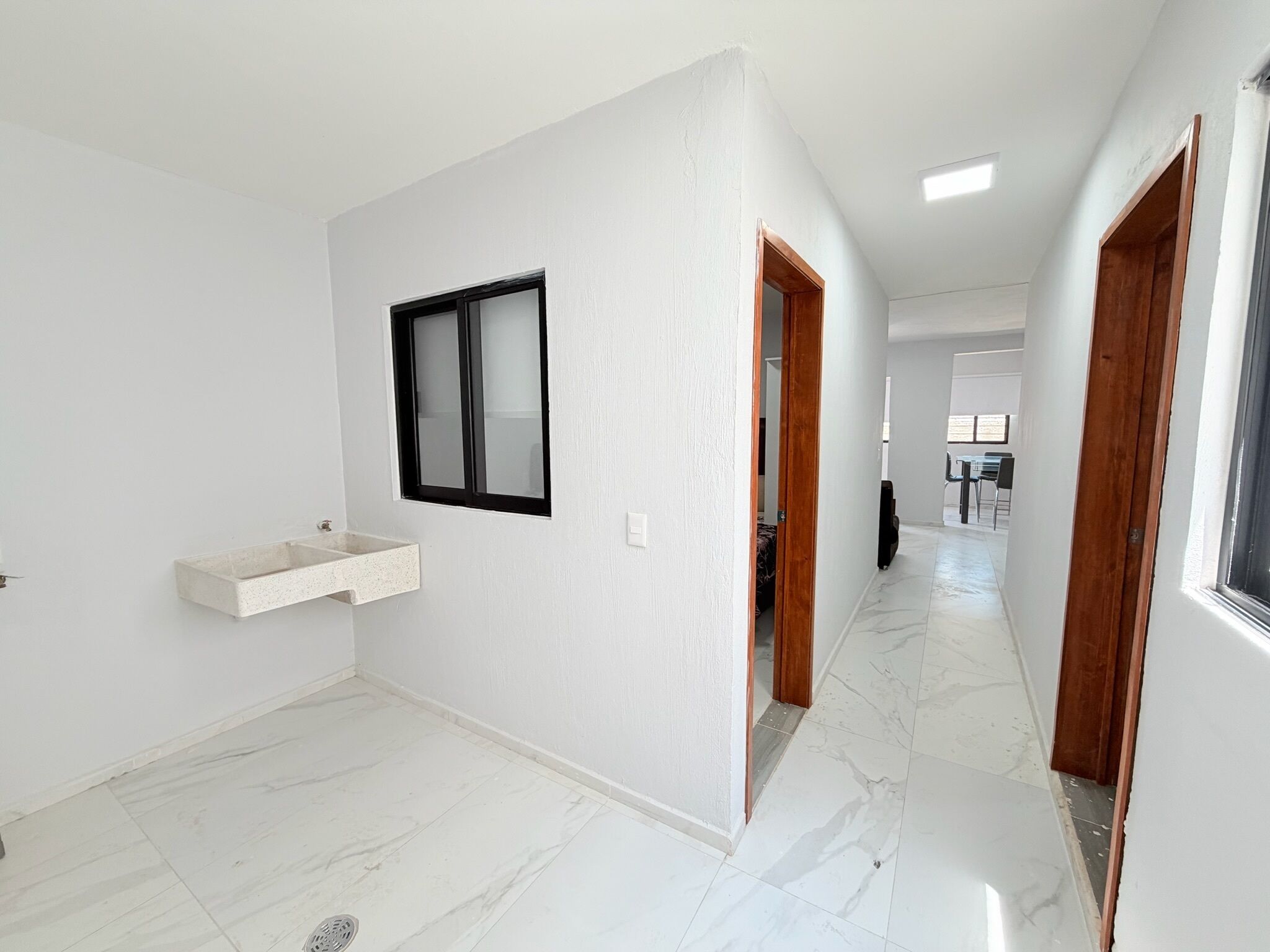 A hallway with a white wall, a window, a wooden door, and a marble floor.