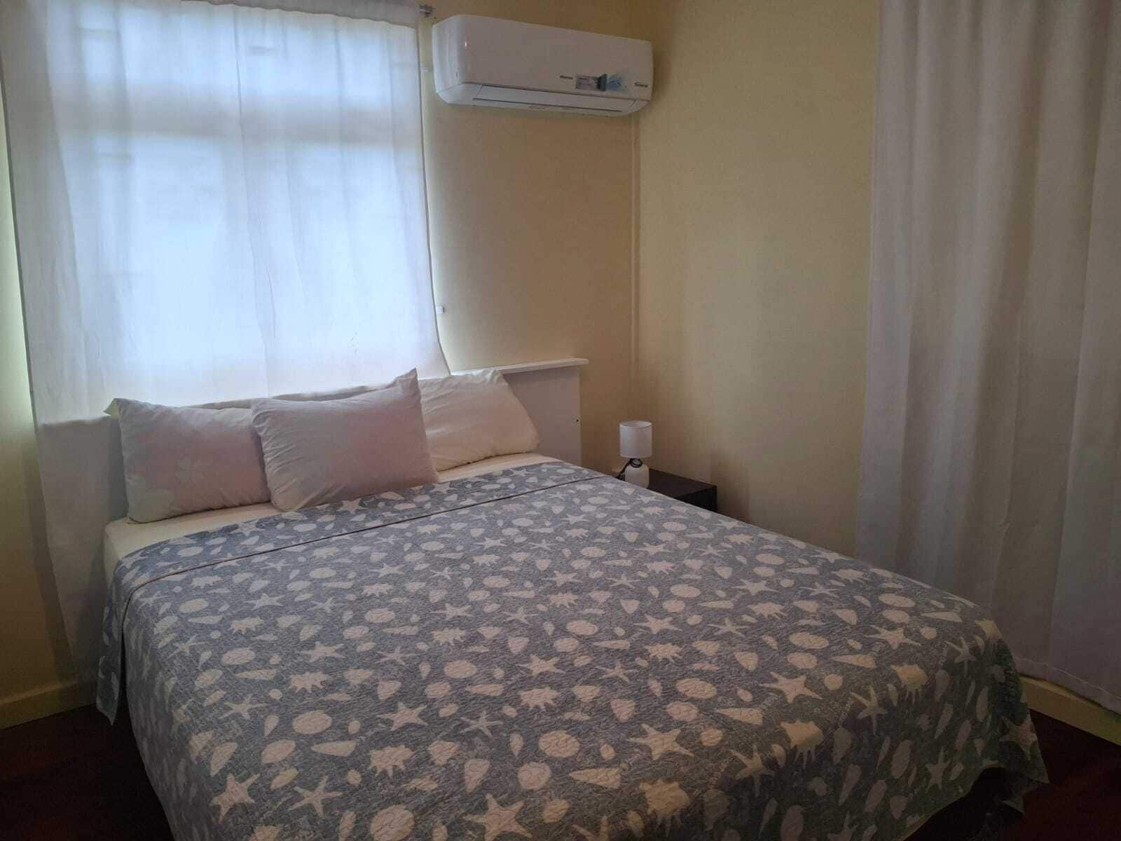 A bedroom with a bed, pillows, a nightstand, an air conditioning unit, and a window with curtains.