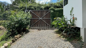 A gravel path leading to a wooden gate, surrounded by greenery and a white building. - Stunning 2BR Home with Fast WiFi & AC – Mountain Views & Lush Nature, Ojochal (Puntarenas Province)