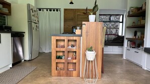A modern kitchen with a built-in wooden cabinet, a white pedestal table with a plant, and a view of a room with a window and curtains. - Stunning 2BR Home with Fast WiFi & AC – Mountain Views & Lush Nature, Ojochal (Puntarenas Province)