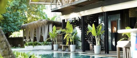 A pool area with potted plants, a thatched structure, and a building with a balcony.