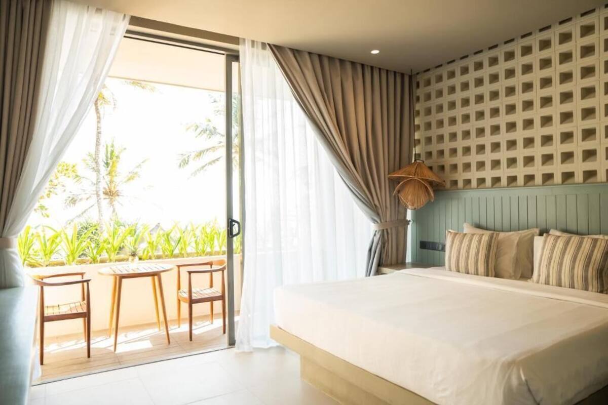 A bedroom with a bed, curtains, a view of palm trees, and a balcony with chairs.