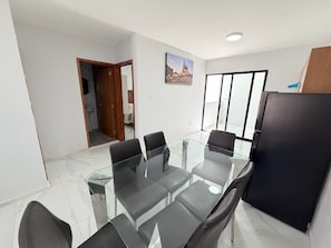 A modern dining area with a glass table and black chairs, a refrigerator, and a large window with a view.