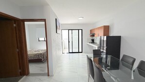 A modern kitchen with a dining area, a bedroom with a bed and window, and a hallway leading to a balcony. - Residencia Gran Coyula Orange 510A (Tonala)