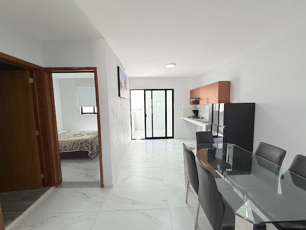 A modern kitchen with a dining area, a bedroom with a bed and window, and a hallway leading to a balcony.