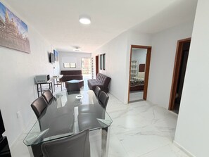 A modern living room with a glass dining table, leather sofas, a TV, and a large window.