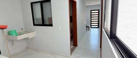 A hallway with a white tiled floor, a window on the right, a door on the left, and a small sink with a green container.