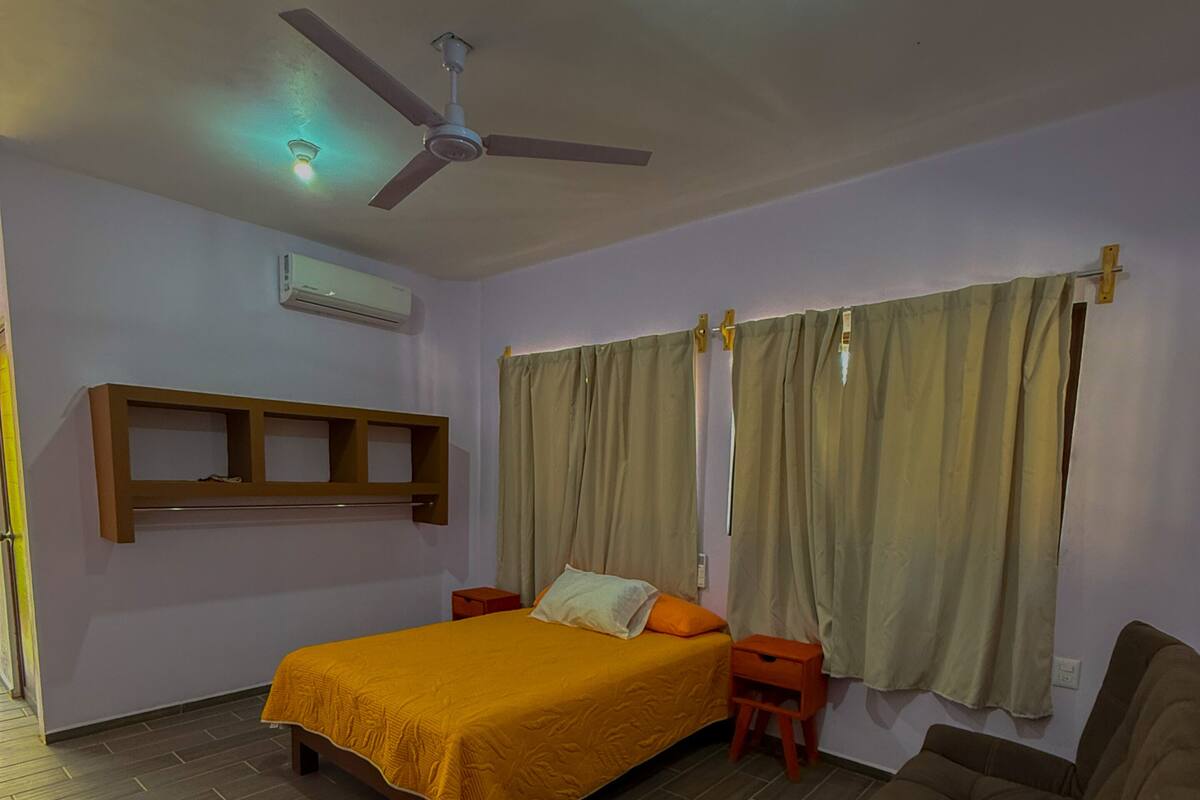 A bedroom with a bed, a ceiling fan, an air conditioning unit, a window with curtains, and a brown sofa.