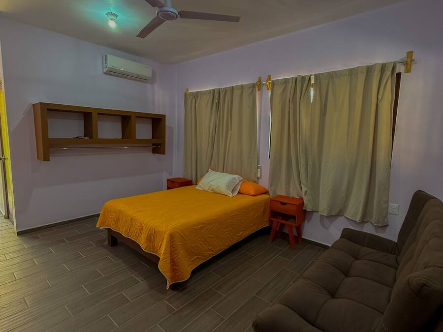 Um quarto com uma cama, um ventilador de teto, um aparelho de ar condicionado, uma janela com cortinas e um sofá marrom.
