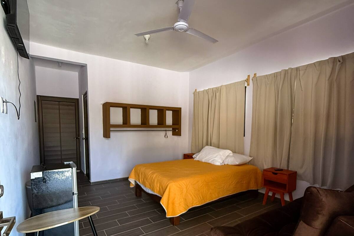 A hotel room with a bed, a chair, a small table, a ceiling fan, and a window with curtains.