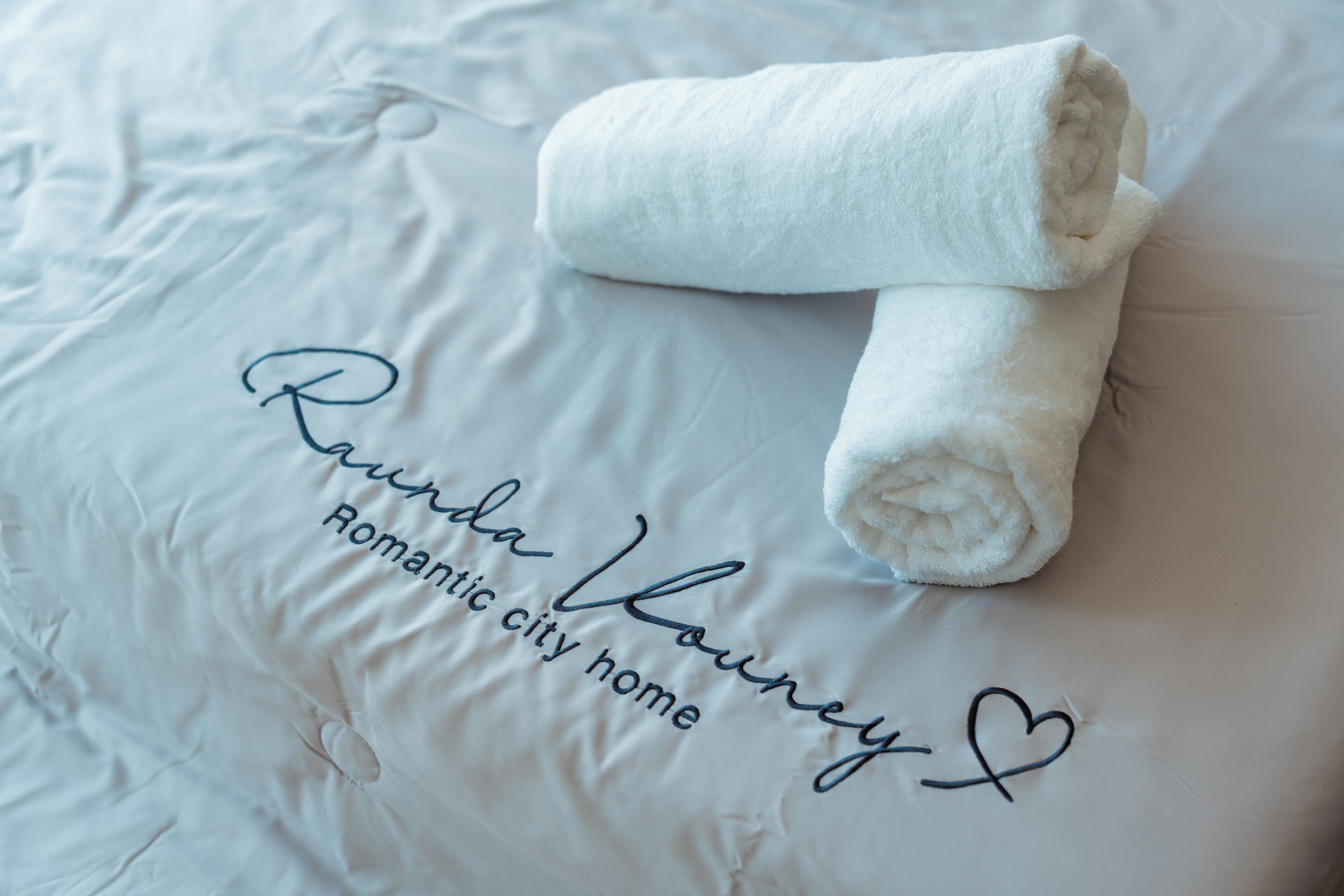 A neatly made bed with a rolled towel and a pillow with 'Raunda Louney' and 'Romantic city home' embroidered on it.