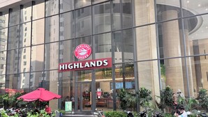 Un bâtiment moderne avec une façade en verre, une enseigne bien visible pour Highland's Coffee, et un espace aménagé avec des tables et des parasols.