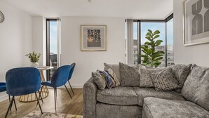 Sala de estar moderna com sofá seccional cinza, cadeiras azuis, mesa de jantar e grandes janelas com vista para a cidade.