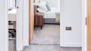 Un pasillo que lleva a un dormitorio con cama, mesita de noche, lámpara y una puerta con interruptor de luz.