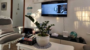 A living room with a television mounted on the wall, a white sofa, a coffee table with a plant, and a bookshelf with decorative items.