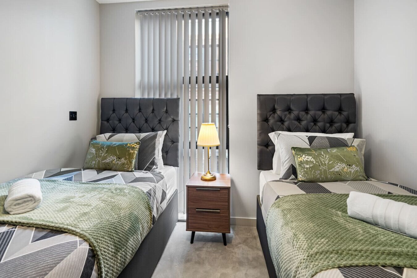 Une chambre d’hôtel avec deux lits, chacun doté d’une tête de lit capitonnée, d’une table de chevet avec une lampe et d’une fenêtre avec des stores verticaux.