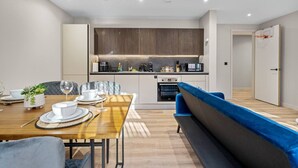 A modern kitchen with built-in appliances, a dining area with a wooden table and blue upholstered bench, and a small potted plant. - Luxury Living | City Views | Spacious | Parking | (West Midlands)