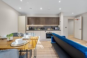 A modern kitchen with built-in appliances, a dining area with a wooden table and blue upholstered bench, and a small potted plant. - Luxury Living | City Views | Spacious | Parking | (West Midlands)