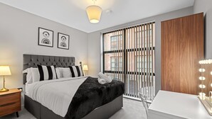 A modern bedroom with a tufted headboard, a large bed, a vanity with lights, and a view through a window with blinds. - Luxury Living | City Views | Spacious | Parking | (West Midlands)