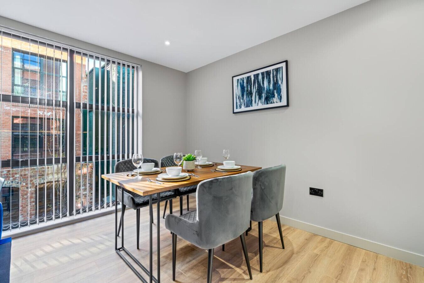 A dining area with a wooden table set for four, grey upholstered chairs, a city view through large windows, and a framed abstract painting on the wall.