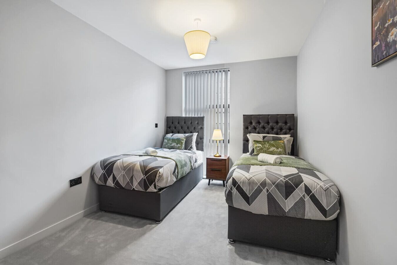 A bedroom with two beds, a nightstand, a lamp, and a window with blinds.
