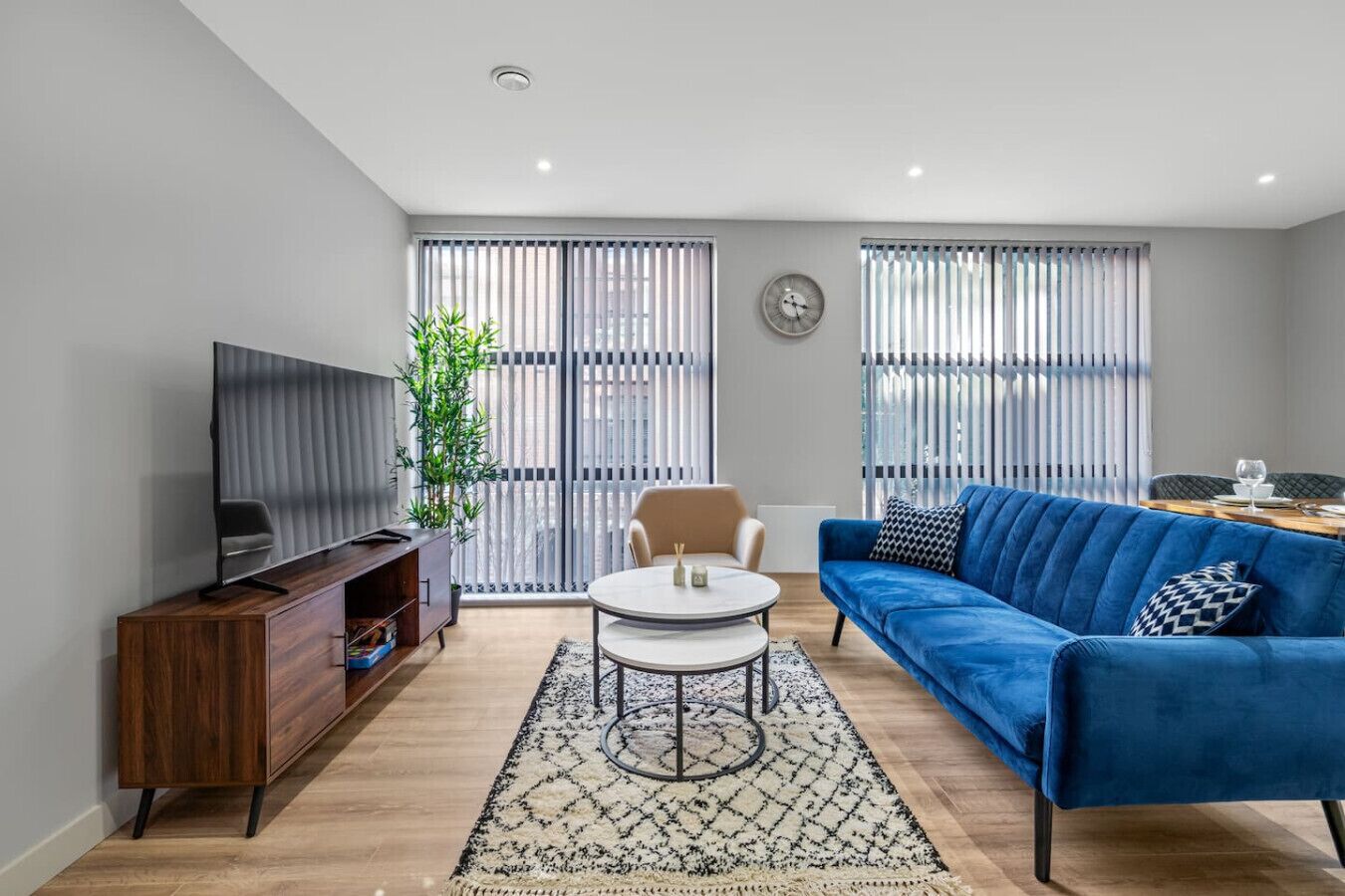 Uma sala de estar moderna com um sofá azul, uma mesa de centro redonda, uma TV em um móvel de madeira e uma janela grande com persianas.