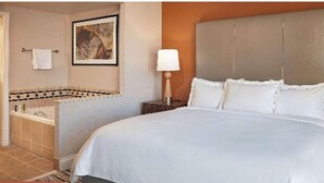Une chambre d’hôtel avec un lit, une table de chevet avec une lampe, une salle de bain avec une douche et un tableau encadré au mur.