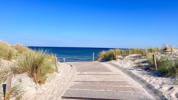 Un sendero de concreto que conduce a la playa, rodeado de dunas de arena y vegetación escasa.