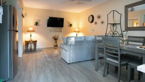 A modern living room with a grey sofa, a dining table with chairs, a flat-screen TV mounted on the wall, and a small side table with a lamp. - Adorable 1-bedroom apartment within 5 minute walk to UF Healthand Proton (Jacksonville)