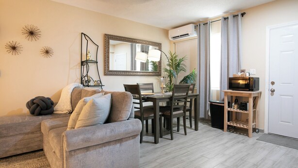 A living room with a sofa, dining table, chairs, a small wooden side table, a potted plant, and a door with an air conditioning unit. - Adorable 1-bedroom apartment within 5 minute walk to UF Healthand Proton (Jacksonville)