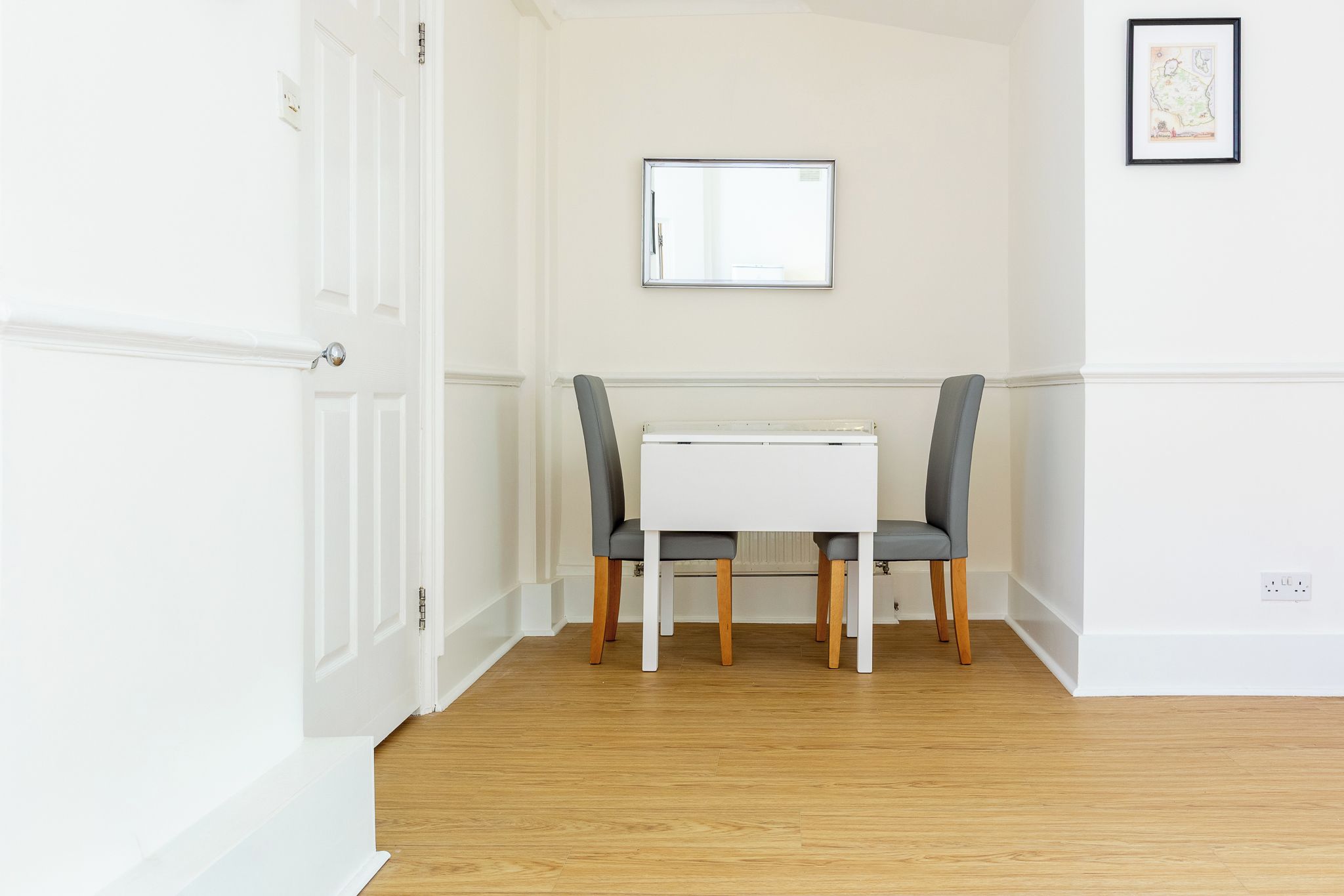 Une pièce avec une porte blanche, une table blanche et deux chaises, et une carte encadrée accrochée au mur.