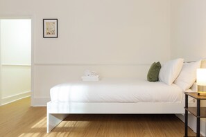 A minimalist bedroom with a white bed, a small bedside table with a lamp, and a framed map on the wall. - Brilliantly Located Studio Flat in Earls Court (Greater London)
