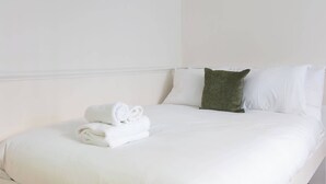 Un lit bien fait, avec des draps blancs et un oreiller vert, une serviette pliée posée à côté, et un mur blanc sobre.