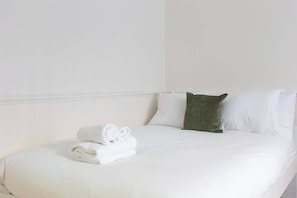 Ein ordentlich bezogenes Bett mit weißen Leinen und einem grünen Kopfkissen, ein gefaltetes Handtuch daneben und eine schlichte weiße Wand.