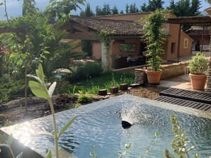 A pool with water splashing, surrounded by potted plants and a building with a tiled roof. - Villa hauteur de Lauris infinity pool not overlooked (Lauris)